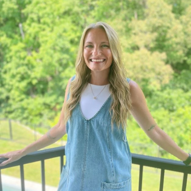 Woman in a denim dress standing on a balcony with a pool and greenery in the background