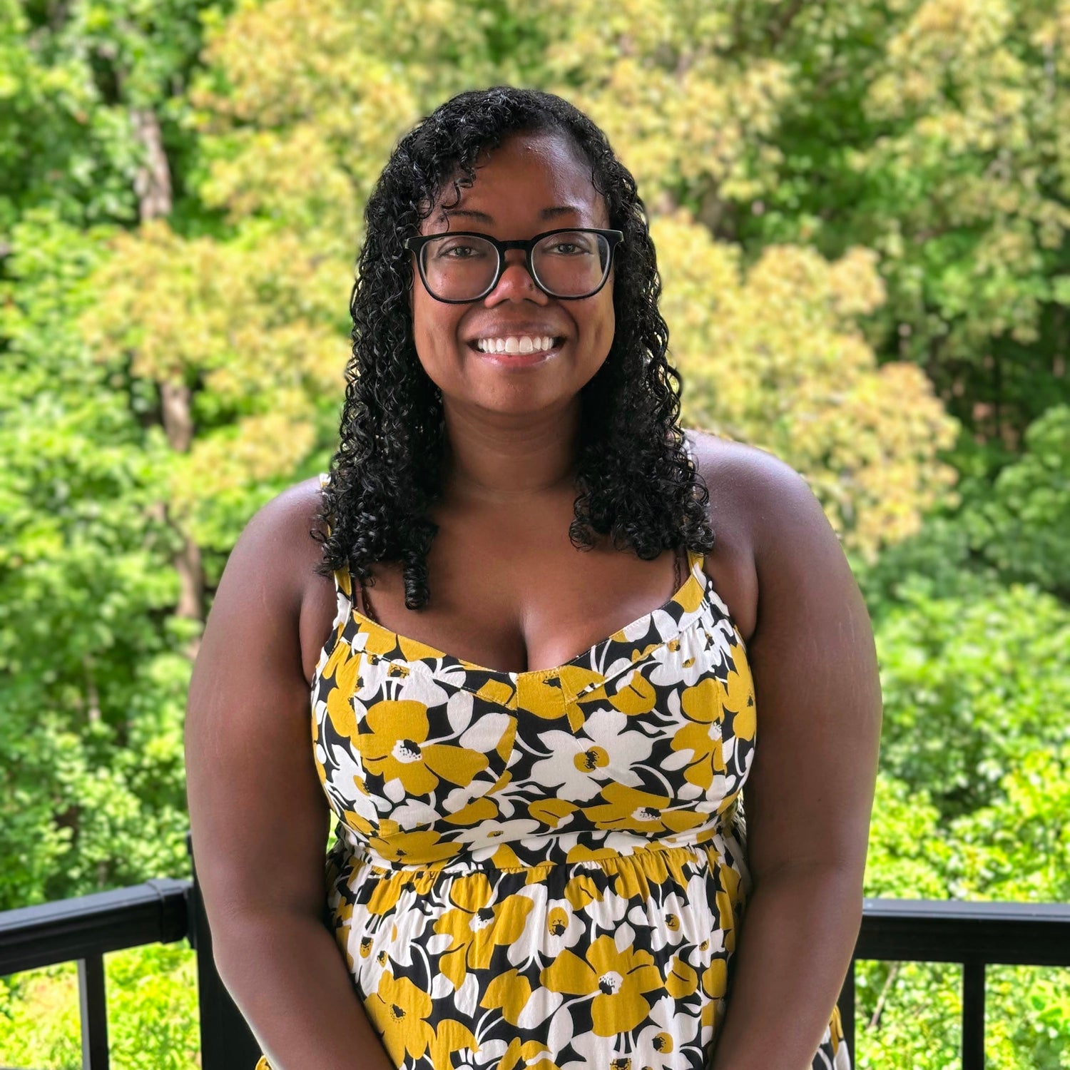 Woman wearing a yellow floral dress standing outdoors with greenery in the background